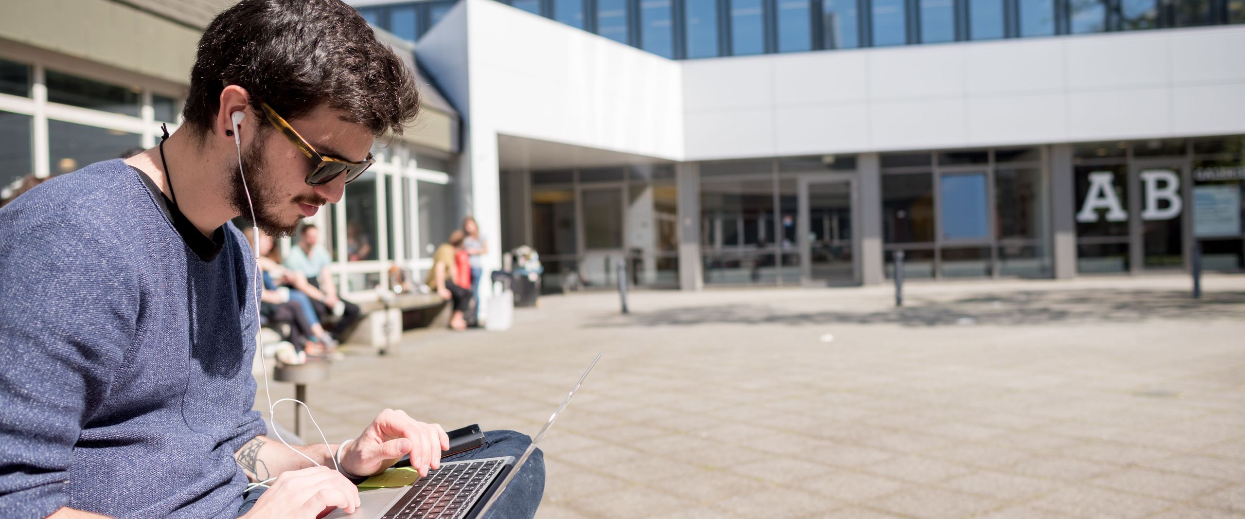 Student sitzt mit Laptop vor dem AB Gebäude draußen