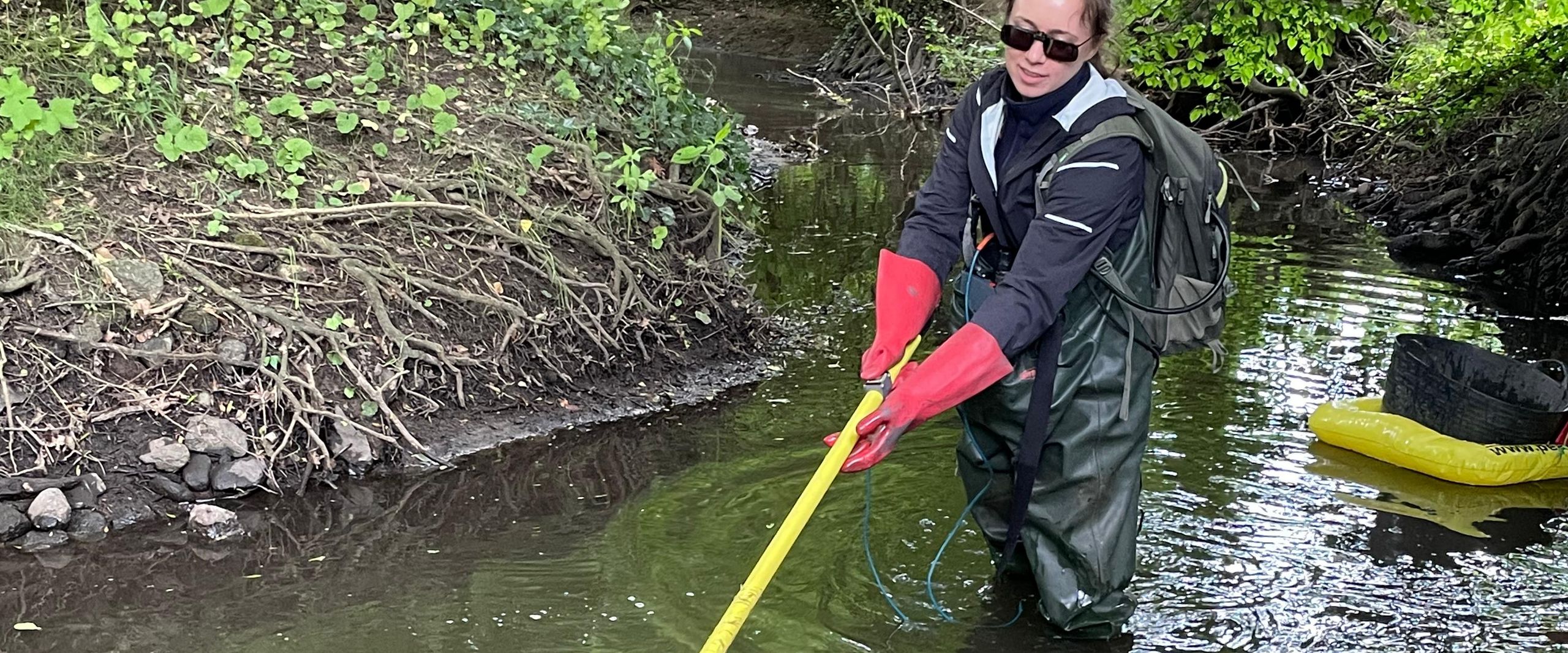 Isabel Tanzberger bei den Gewässeruntersuchungen Eine Frau mit Rucksack steht im wasserdichten Anzug in einem kleinen Wasserlauf. Das Wasser reicht bis zu den Oberschenkeln. Sie taucht eine gelbe Stange ins Wasser, eventuell ein Fischköcher. Im Hintergrund schwimmt ihre weitere Ausrüstung auf einer Luftmatratze.