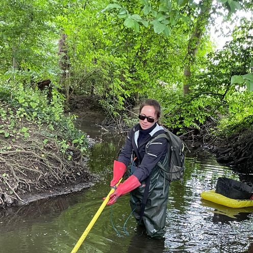 Eine Frau mit Rucksack steht im wasserdichten Anzug in einem kleinen Wasserlauf. Das Wasser reicht bis zu den Oberschenkeln. Sie taucht eine gelbe Stange ins Wasser, eventuell ein Fischköcher. Im Hintergrund schwimmt ihre weitere Ausrüstung auf einer Luftmatratze.