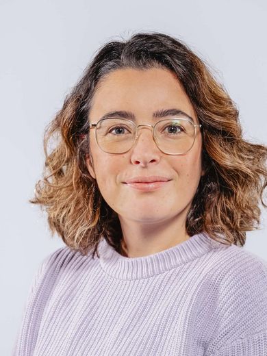 Auf dem Bild ist Annika Müller zu sehen. Sie hat schulterlanges, lockiges, braunes Haar, trägt eine Brille und einen fliederfarbenen Pullover an.