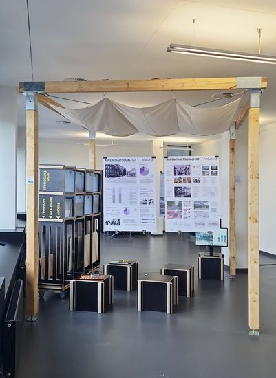 Ausstellungspavillon im Innenraum mit Holzrahmen und darüber gespanntem Stoffdach. Im Hintergrund hängen große Plakate mit Informationen und Diagrammen zum Thema Aufenthaltsqualität. Im Vordergrund stehen schwarze Sitzwürfel und seitlich ein Monitor.