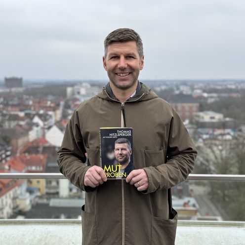 Mann h&auml;lt Buch und lacht in die Kamera