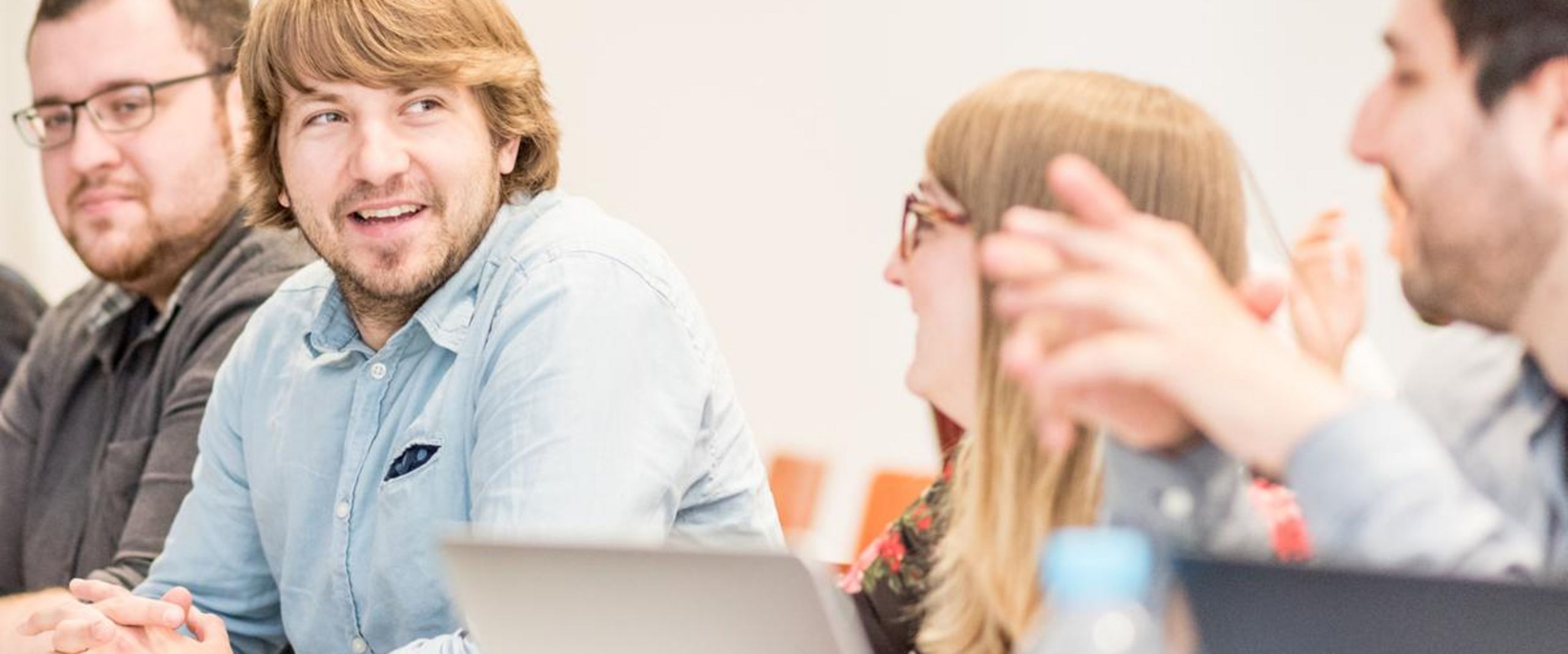 Vier Studierende sitzen gemeinsam an einem Tisch, unterhalten sich und arbeiten mit Laptops. Die Stimmung wirkt konzentriert und freundlich.