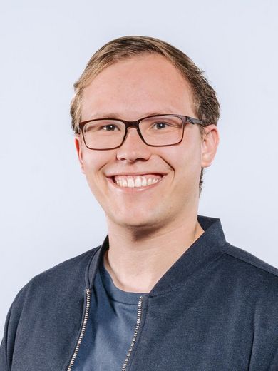 Auf dem Bild ist Tim Gust zusehen. Er hat blonde Haare und trägt eine Brille. Er hat ein blaues T-Shirt unter einer dunkelblauen Jacke an. 