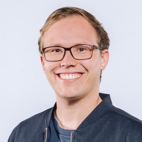 Auf dem Bild ist Tim Gust zusehen. Er hat blonde Haare und trägt eine Brille. Er hat ein blaues T-Shirt unter einer dunkelblauen Jacke an. 