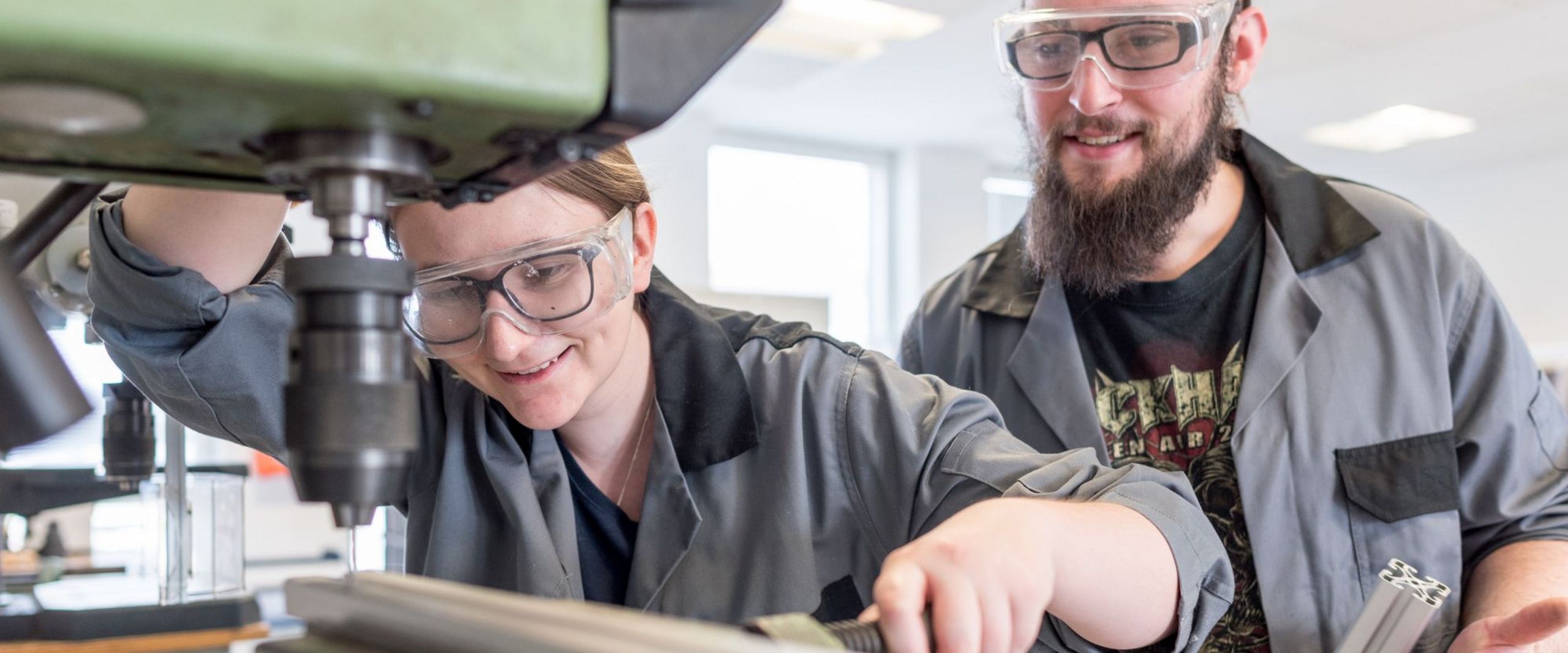 Eine Studentin spannt eine Metallstange in eine Maschine ein, während ein zweiter Student ihr dabei zusieht.