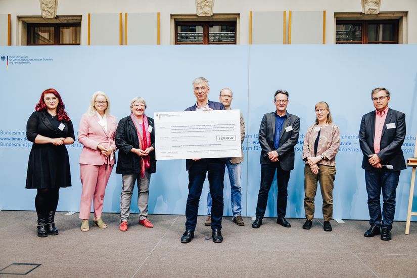 Gruppenbild mit Vertreter:innen des Bundestags, des Bundesumweltministeriums und des Projekts "Smart Recycling Up". Staatssekretär Stefan Tidow präsentiert einen großen Scheck.