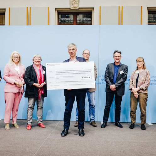 Gruppenbild mit Vertreter:innen des Bundestags, des Bundesumweltministeriums und des Projekts "Smart Recycling Up". Staatssekret&auml;r Stefan Tidow pr&auml;sentiert einen gro&szlig;en Scheck.
