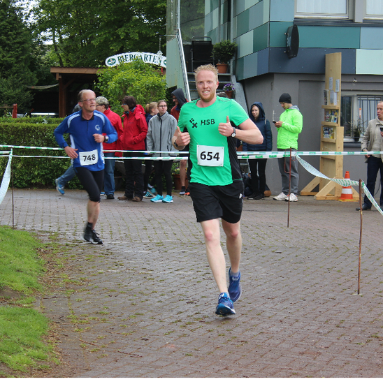 Zwei L&auml;ufer laufen die Strecke entlang. Im Hintergrund steht ein Geb&auml;ude und mehrere Personen.