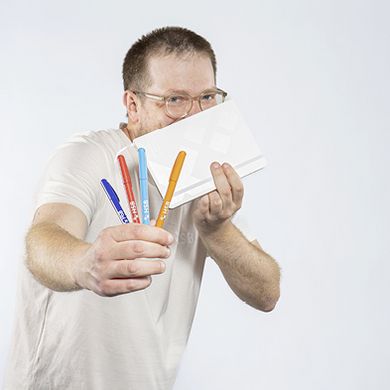 Ein Student h&auml;lt mit der linken Hand ein wei&szlig;es Notizbuch mit HSB-Logo vor sein Gesicht, sodass sein Mund verdeckt ist. Mit der rechten Hand h&auml;lt er 4 bunte Kugelschreiber mit HSB-Logo in die Kamera.