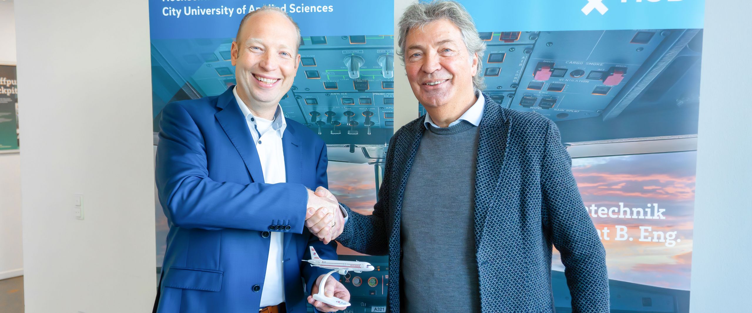 Zwei Männer schütteln sich die Hand. Der linke hält ein kleines Modellflugzeug.