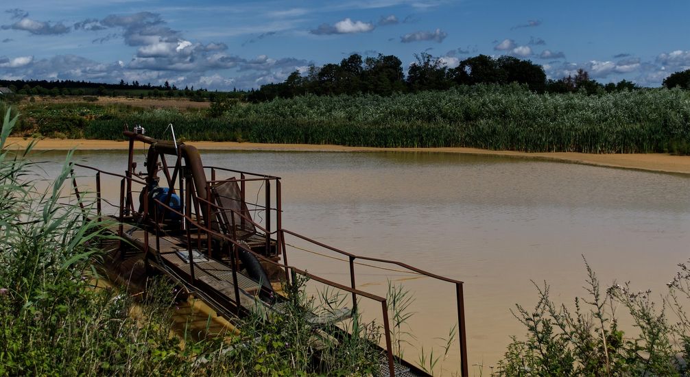 Ein Baggersee im Sommer mit gelblich-trübem, sandigem Wasser, einer alten, verrosteten Förderanlage im Vordergrund und grünem Uferbewuchs. Darüber blauer Himmel.