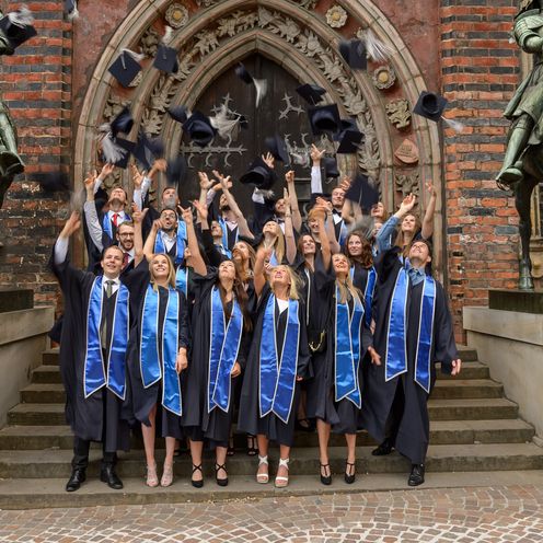 Absolvent:innen des Dualen Studiengangs Betriebswirtschaft im Talar werfen ihre H&uuml;te vor dem Bremer Rathaus in die Luft.