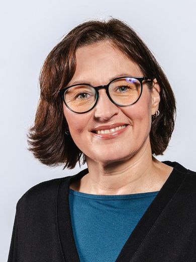 Auf dem Foto ist Alexandra Gutherz zusehen. Sie hat dunkelbraunes kinnlanges Haar. Sie trägt eine Brille und ein blaues Shirt unter einem schwarzen Cadigan.