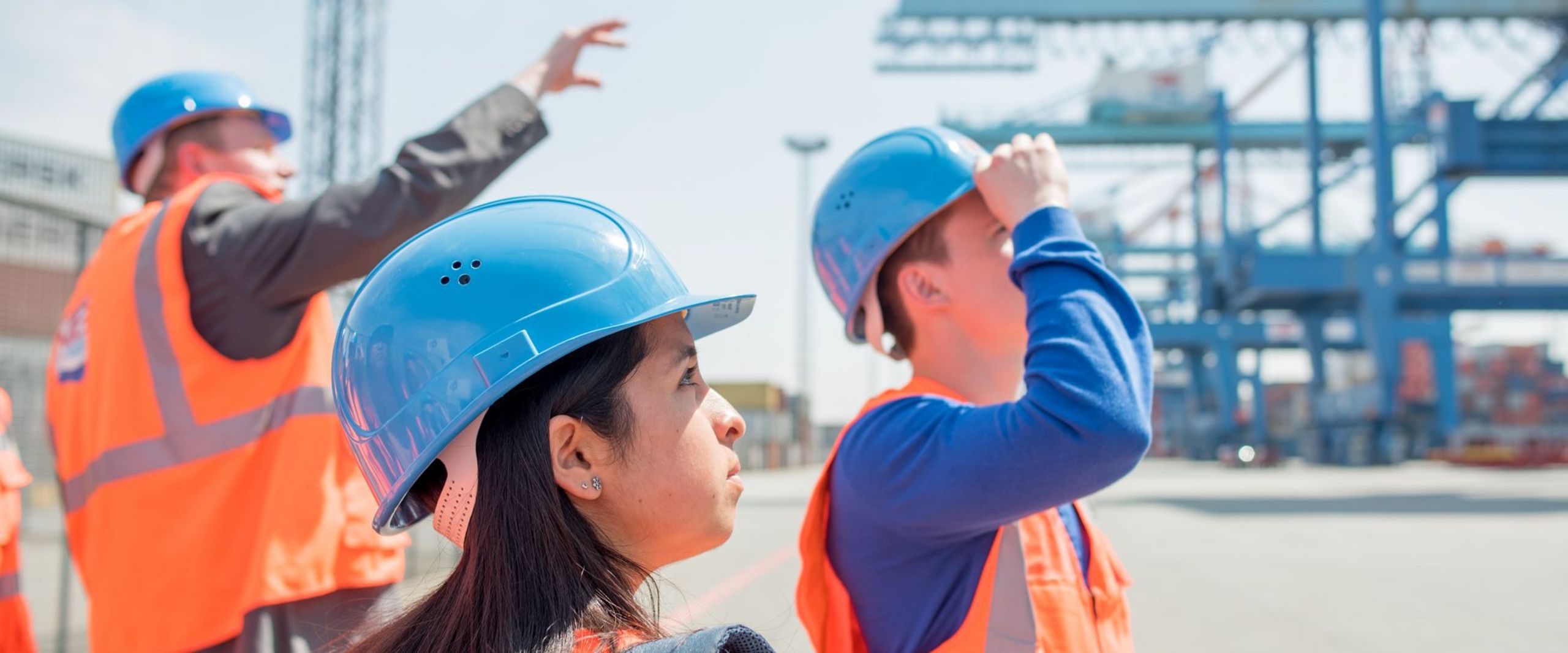 Ein Beschäftigter führt Studierende in Warnwesten und Schutzhelmen durch ein Containerterminal.