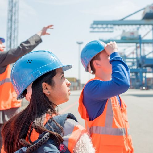 Ein Besch&auml;ftigter f&uuml;hrt Studierende in Warnwesten und Schutzhelmen durch ein Containerterminal.