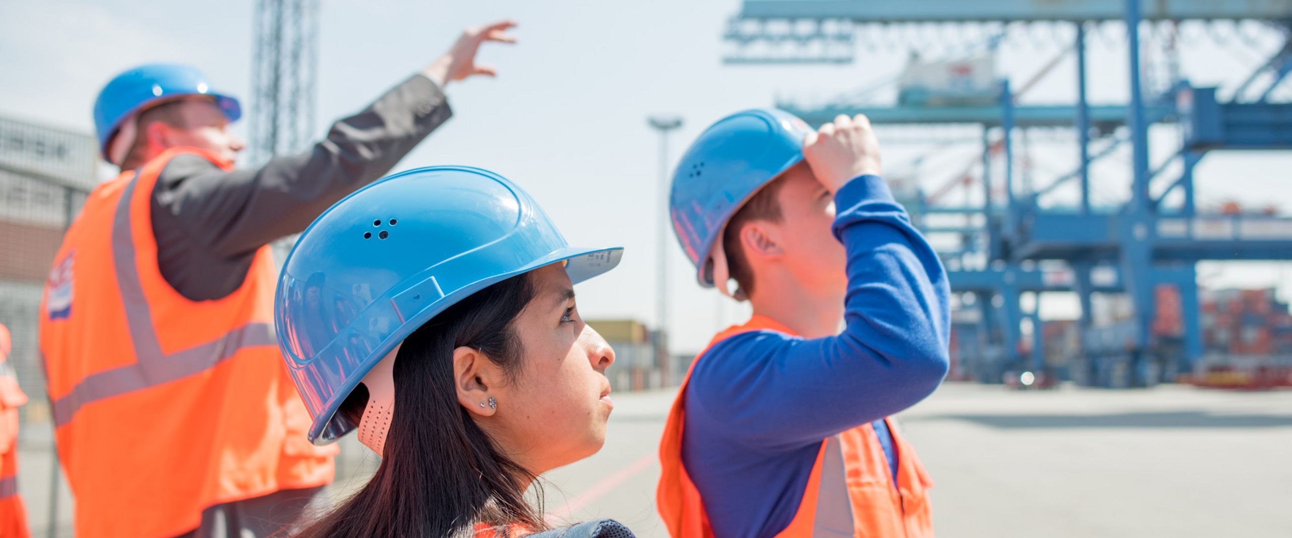 Ein Beschäftigter führt Studierende in Warnwesten und Schutzhelmen durch ein Containerterminal.