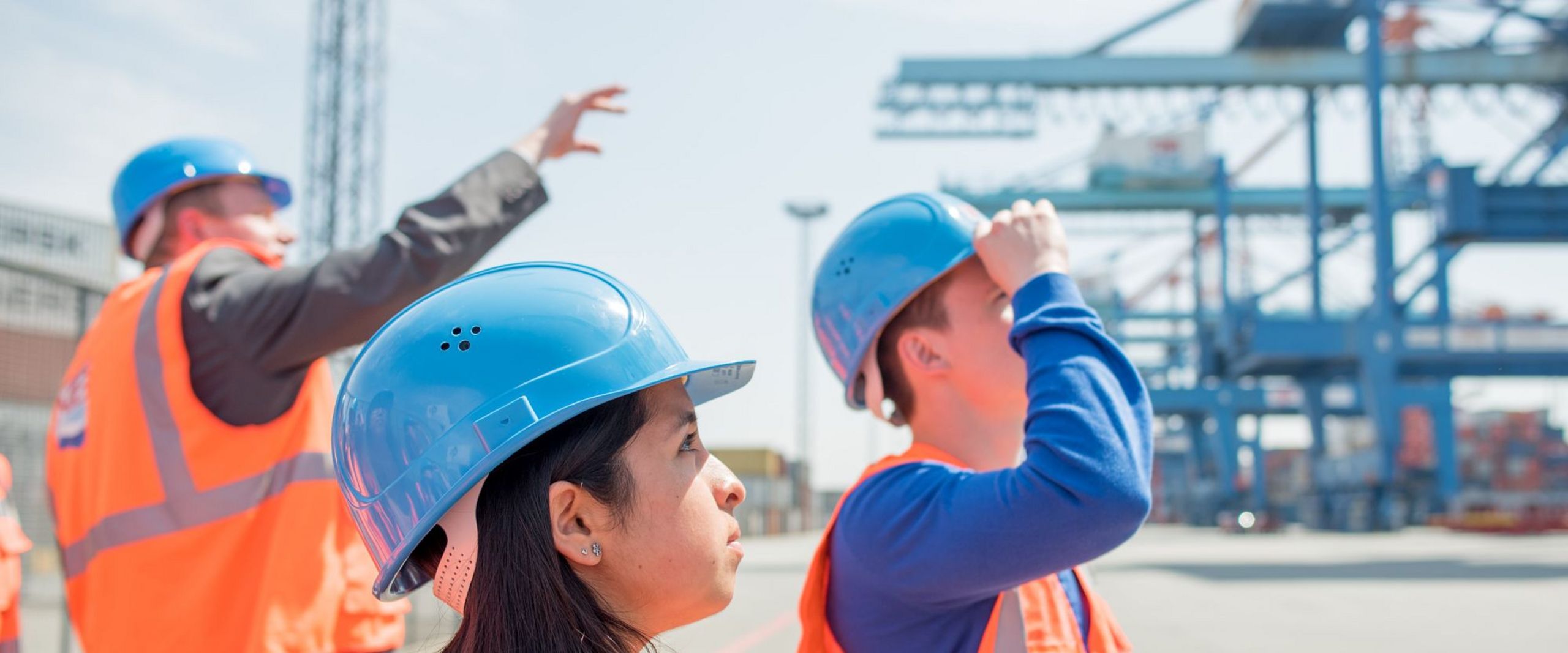 Ein Beschäftigter führt Studierende in Warnwesten und Schutzhelmen durch ein Containerterminal.