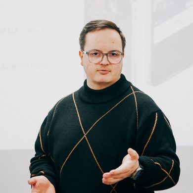 Auf dem Bild ist ein junger Mann mit kurzen braunen Haaren zusehen. Er trägt eine gründliche Brille und einen schwarzen Pullover mit einem dünnen orangenen X, das sich über den Oberkörper streckt. 