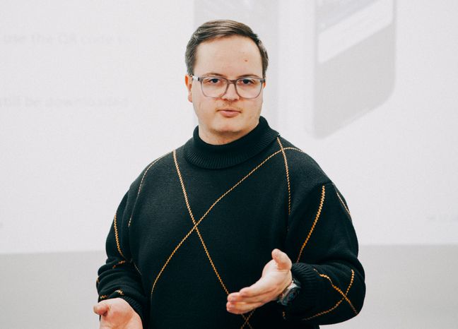 Sebastian Mertins Auf dem Bild ist ein junger Mann mit kurzen braunen Haaren zusehen. Er trägt eine gründliche Brille und einen schwarzen Pullover mit einem dünnen orangenen X, das sich über den Oberkörper streckt.