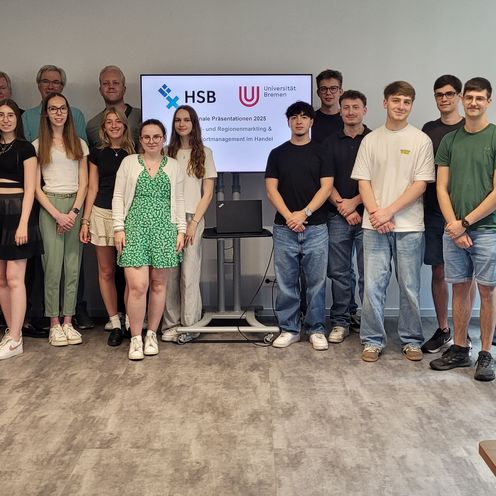Das Bild zeigt eine Gruppe von circa zwanwig Studierenden sowie sieben weiteren Gästen und Dozenten, die vor einer Wand stehen. In der Mitte ist ein TV-Monitor zu sehen, auf dem die Logos der Hochschule Bremen und der Universität Bremen zu erkennen sind.