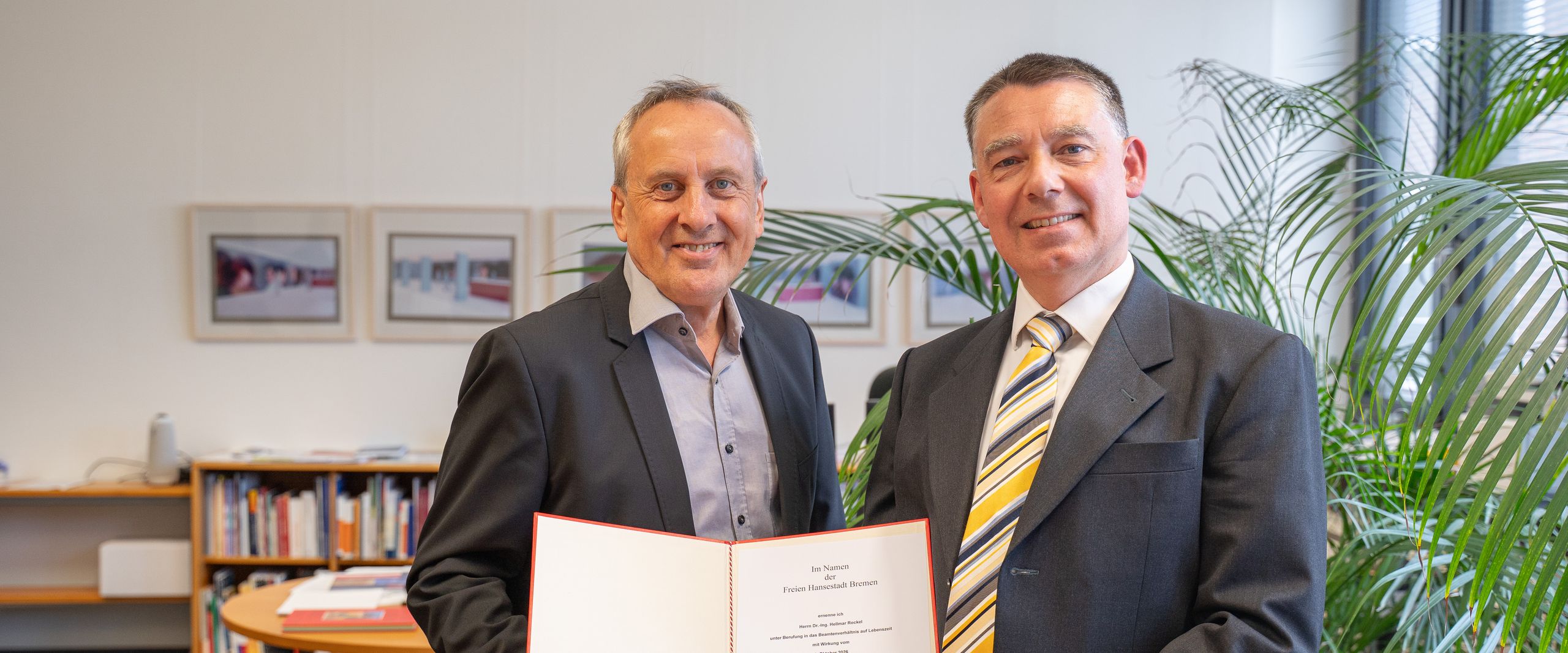 202608_Hellmar Rockel und Rektor Zwie Männer halten gemeinsam eine Urkunde und lächeln in die Kamera