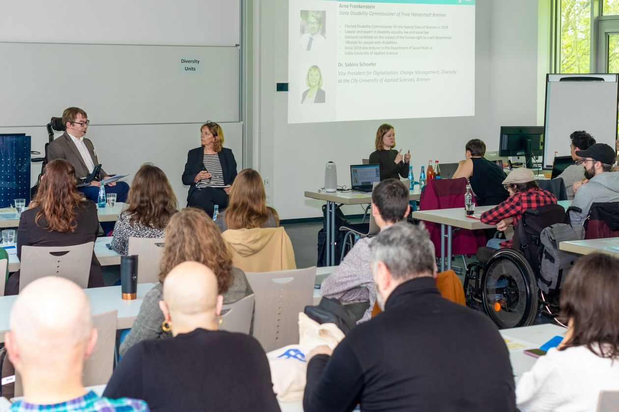 Seminarraum mit Zuhörer:innen. Vorne sind ein mann im Rollstuhl und eine Frau im Gespräch. Rechts übersetzt eine Frau in Gebärdensprache