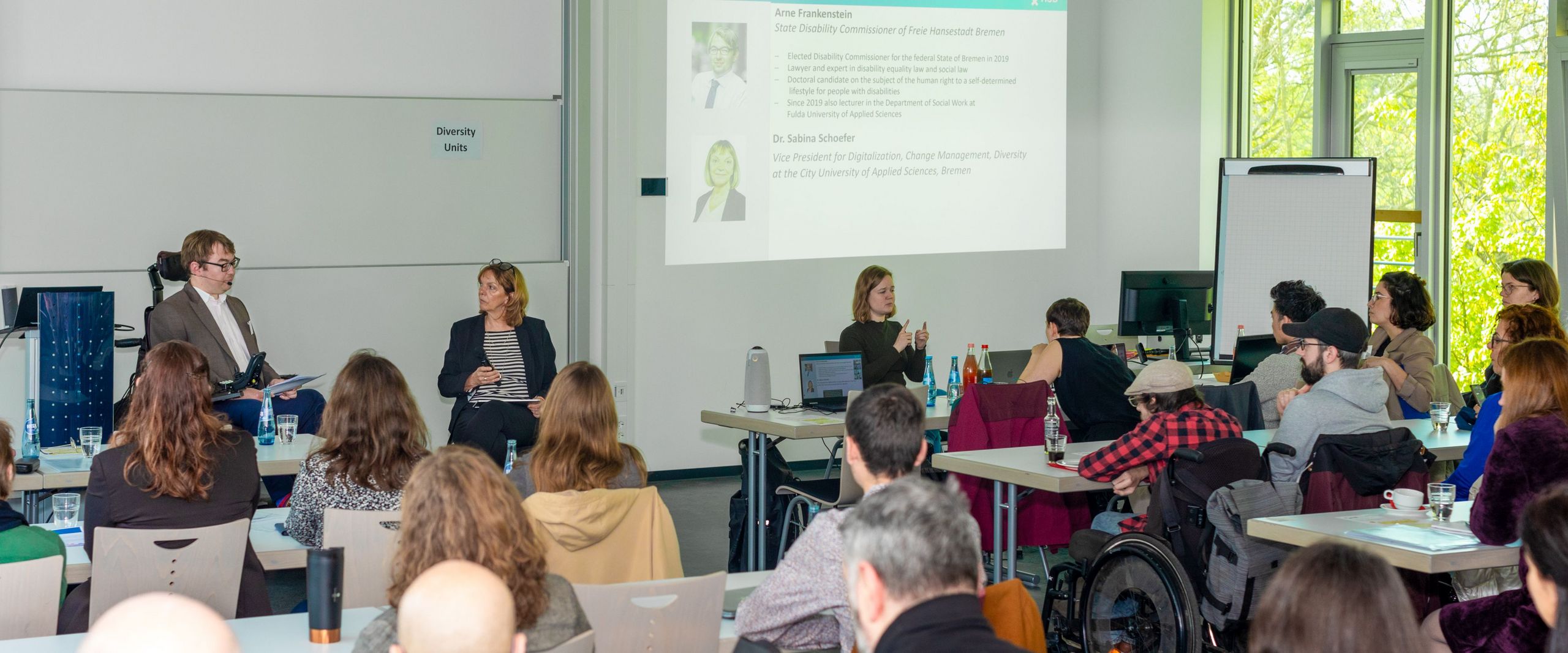 Seminar room with listeners. In front, a man in a wheelchair and a woman are talking. On the right, a woman translates into sign language