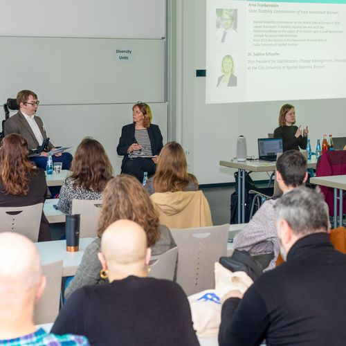 Seminar room with listeners. In front, a man in a wheelchair and a woman are talking. On the right, a woman translates into sign language