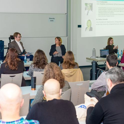 Seminarraum mit Zuh&ouml;rer:innen. Vorne sind ein mann im Rollstuhl und eine Frau im Gespr&auml;ch. Rechts &uuml;bersetzt eine Frau in Geb&auml;rdensprache