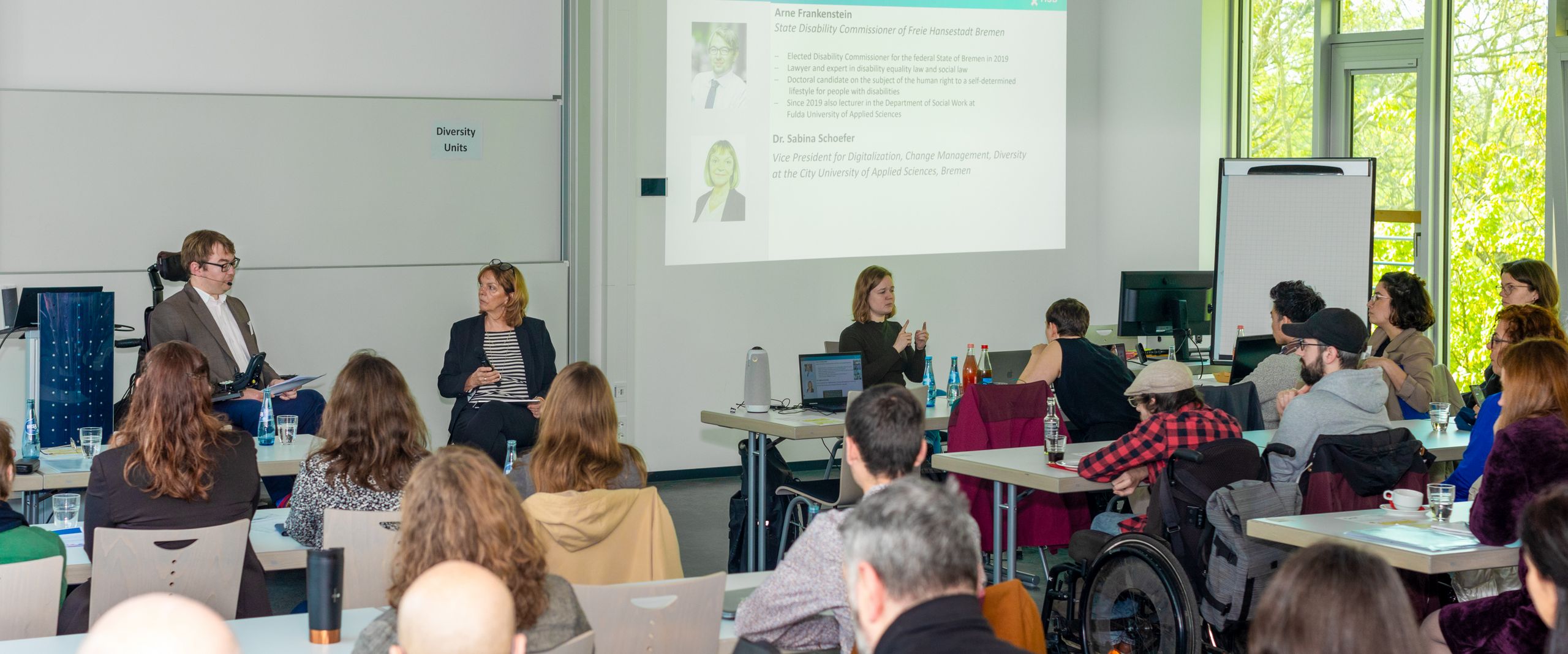 Seminarraum mit Zuhörer:innen. Vorne sind ein mann im Rollstuhl und eine Frau im Gespräch. Rechts übersetzt eine Frau in Gebärdensprache