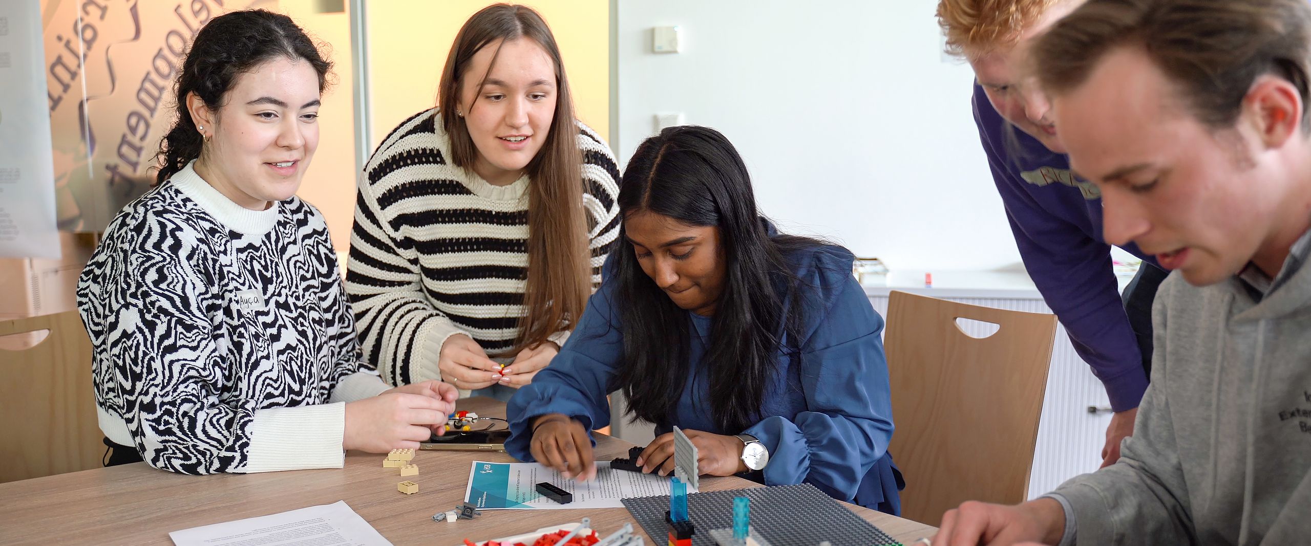 Auf dem Bild ist eine fünfköpfige Gruppe aus drei Studentinnen und zwei Studenten zu sehen. Sie sitzen gemeinsam an einem Tisch. Vor ihnen auf dem Tisch liegen Legosteine. 