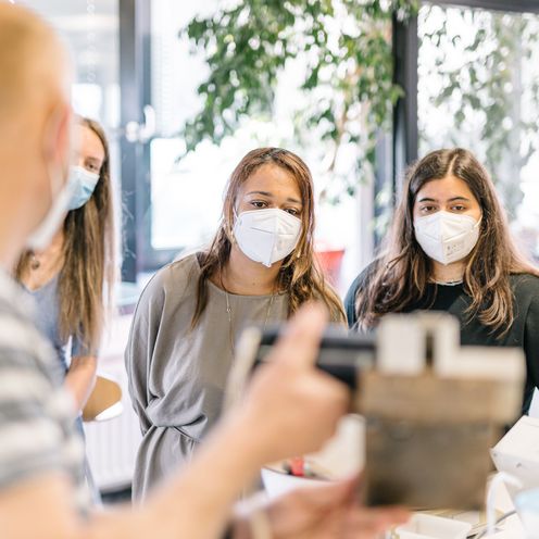 Zwei Studierende mit Masken h&ouml;ren dem Professor zu.