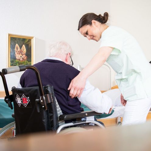 Eine Altenpflegerin hilft einem &auml;lteren Mann, sich in einen Rollstuhl zu setzen.