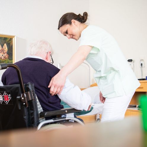 Eine Altenpflegerin hilft einem &auml;lteren Mann, sich in einen Rollstuhl zu setzen.