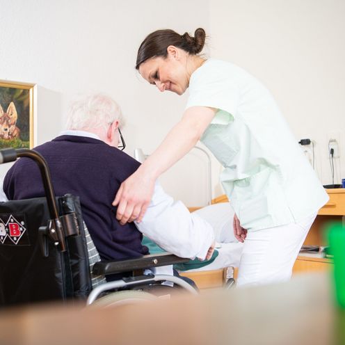 Eine Altenpflegerin hilft einem &auml;lteren Mann, sich in einen Rollstuhl zu setzen.
