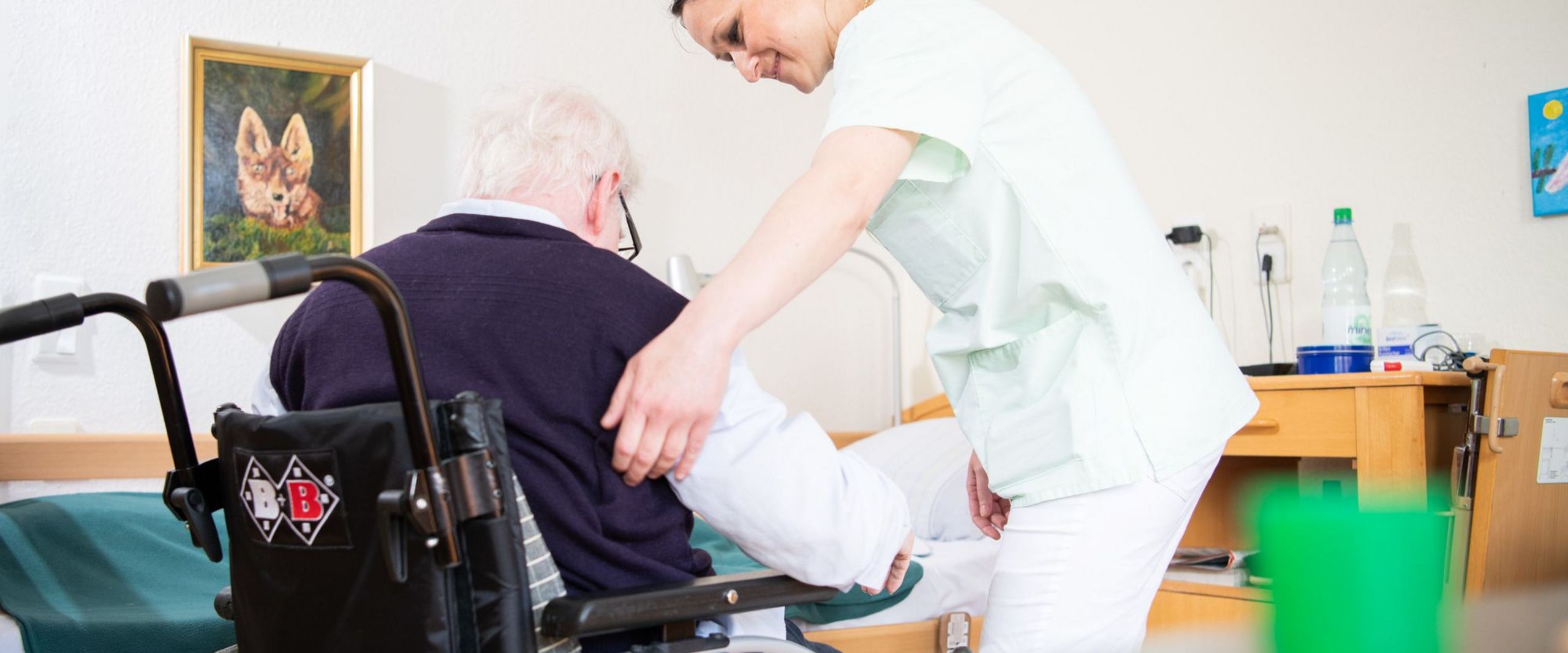 Eine Altenpflegerin hilft einem älteren Mann, sich in einen Rollstuhl zu setzen.