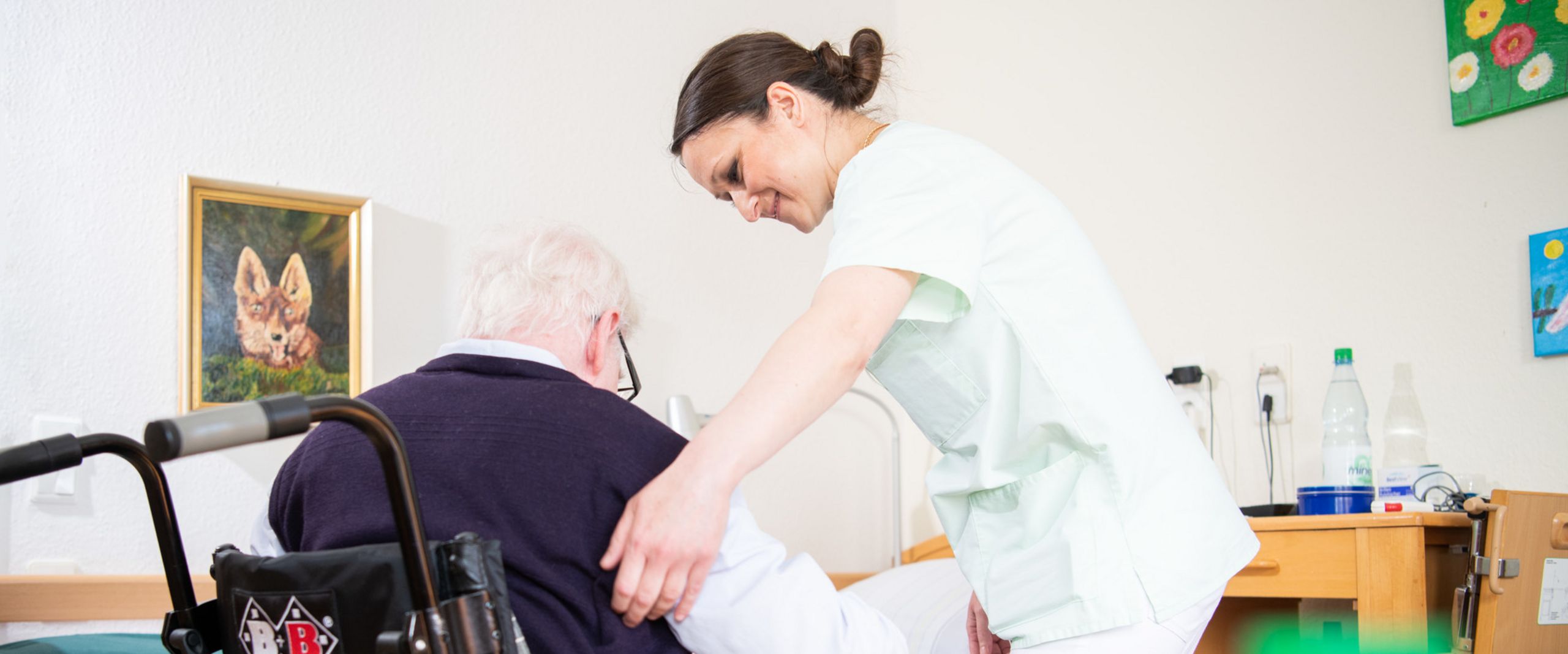 Eine Altenpflegerin hilft einem älteren Mann, sich in einen Rollstuhl zu setzen.