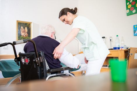 Eine Altenpflegerin hilft einem älteren Mann, sich in einen Rollstuhl zu setzen.
