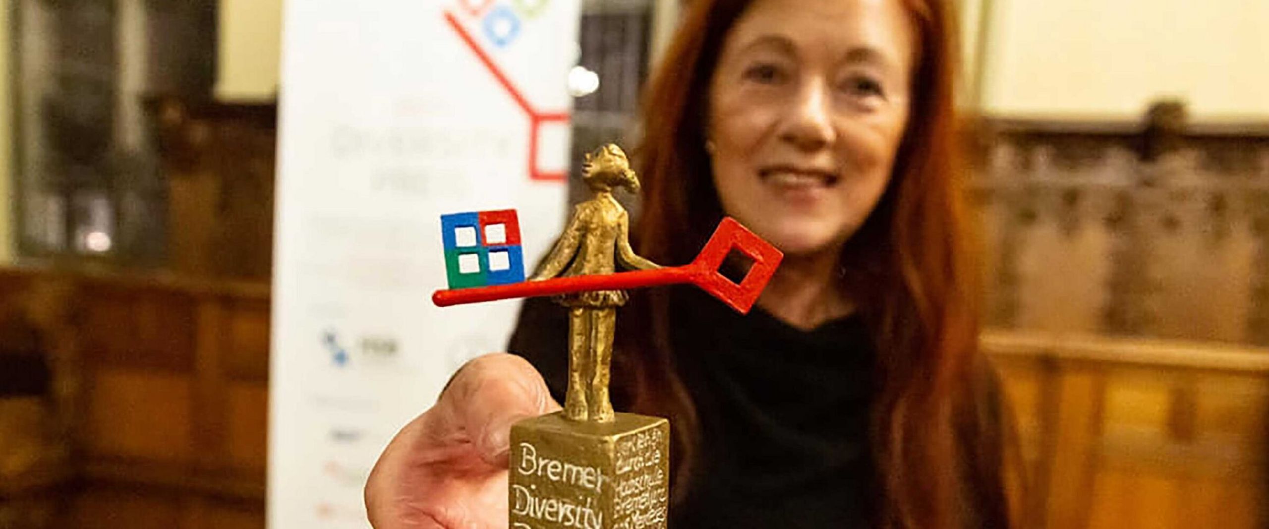Frau hält Skulptur in die Kamera. Im Hintergrund sieht man ein Roll-up