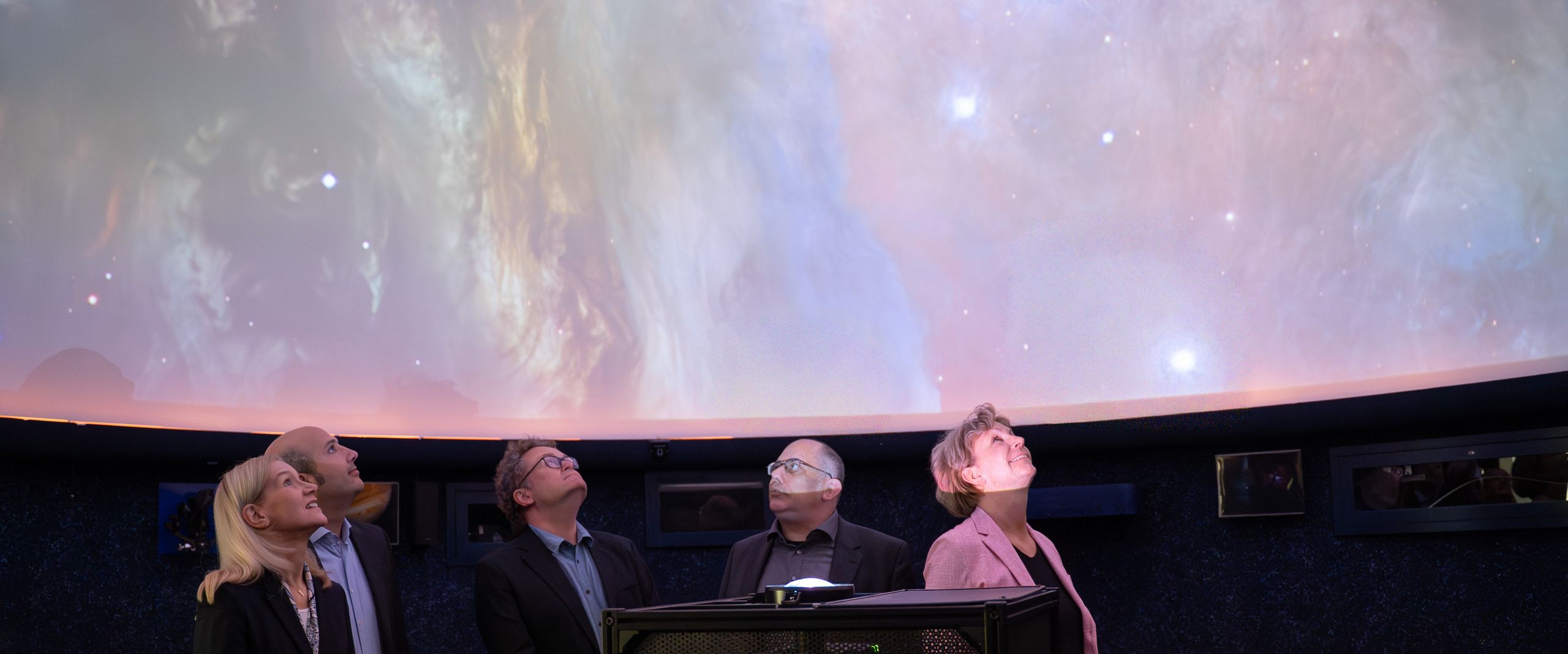 Drei Männer und zwei Frauen blicken im Dunkeln in den Sternenhimmel in einem Planetarium.