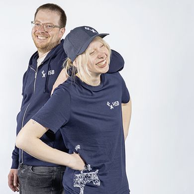 Ein Student in einem dunkelblauen Zip-Hoodie mit HSB-Logo lehnt mit seinem Arm auf der Schulter einer Studentin, die ein dunkelblaues T-Shirt und eine dunkelblaue Baseballcap mit HSB-Logo tr&auml;gt.