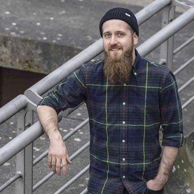 Auf dem Bild ist Alexander Anschütz zusehen. Er trägt eine dunkelblaue Wollmütze, ein grob kariertes dunkelblaues Hemd und einen Bart. Er lehnt an einem Metallgeländer. 