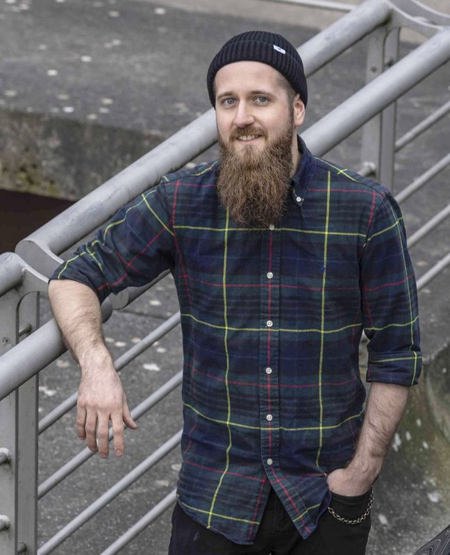 Alexander Anschütz Auf dem Bild ist Alexander Anschütz zusehen. Er trägt eine dunkelblaue Wollmütze, ein grob kariertes dunkelblaues Hemd und einen Bart. Er lehnt an einem Metallgeländer.