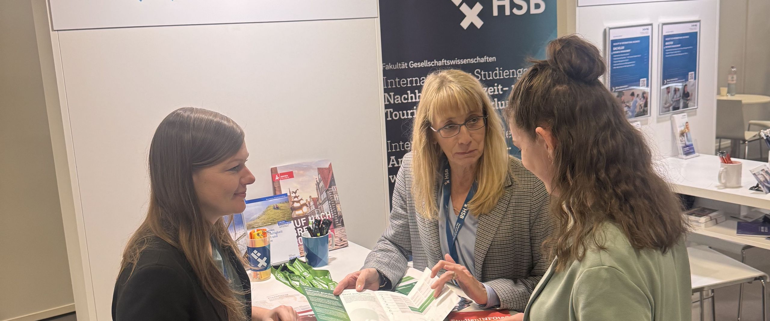 Auf dem Bild ist der Messestand der Hochschule Bremen auf der Internationalen Tourismusbörse zusehen. An dem Stand stehen drei Frauen, die sich unterhalten. 