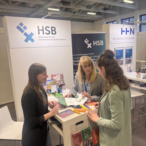 Auf dem Bild ist der Messestand der Hochschule Bremen auf der Internationalen Tourismusb&ouml;rse zusehen. An dem Stand stehen drei Frauen, die sich unterhalten. 