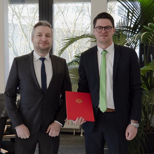 Zwei M&auml;nner l&auml;cheln in die kamera. Der rechte zeigt eine Urkunde.