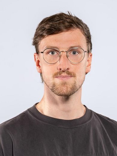 Auf dem Bild ist Jonas Baumann zusehen. Er hat kurzes braunes Haar und einen leichten Bart. Er tr&auml;gt eine Brille und ein dunkelgraues T-Shirt. 