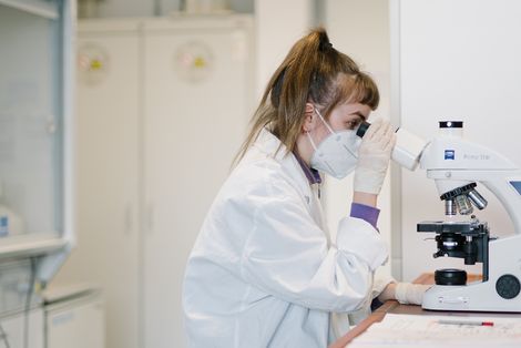 Eine Studentin mit Maske schaut sich durch ein Mikroskope einen Stoff an.