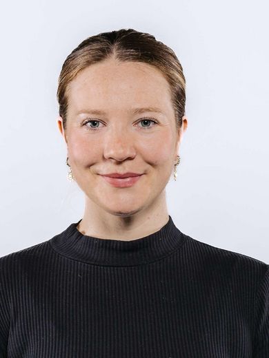 Auf dem Bild ist Greta Kottwitz zusehen. Sie hat ihre blonden Haare in einem tiefen Zopf zusammengebunden. Sie trägt ein hochgeschlossenes schwarzes Shirt. 